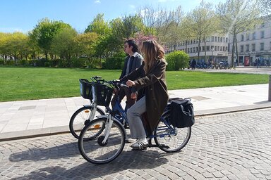 Deux cyclistes, un homme et une femme, circulant côte à côte sur les pavés d'une place en plein soleil printanier