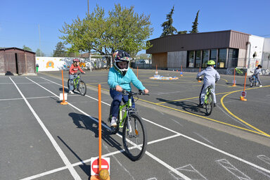 Enfants casqués manœuvrant à vélo sur un parcours balisé de cônes et marquages au sol dans une cour d'école