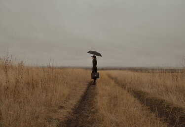une femme dans un champ. Elle s'abrite sous un parapluie et tient une sacoche à la main.