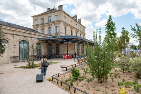 Parvis réaménagé avec arbres, végétation et bancs en bois, un voyageur tirant une valise vers l'entrée de la gare