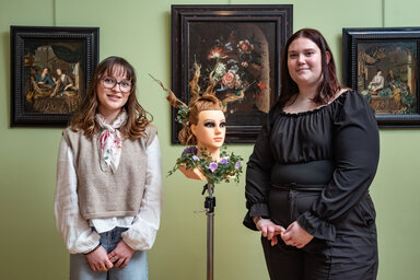 Deux apprenties coiffeuses posent dans une salle du musée encadrant une tête à coiffer ornée d'un chignon fleuri élaboré