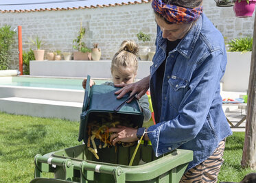 Adulte et enfant déposant des épluchures dans un bac vert à biodéchets dans un jardin