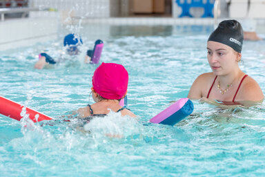 Monitrice en bonnet Arena accompagnant un enfant avec frite et planche dans un bassin couvert